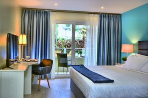 A hotel room with a bed, desk, chair, and a view of a balcony with plants.