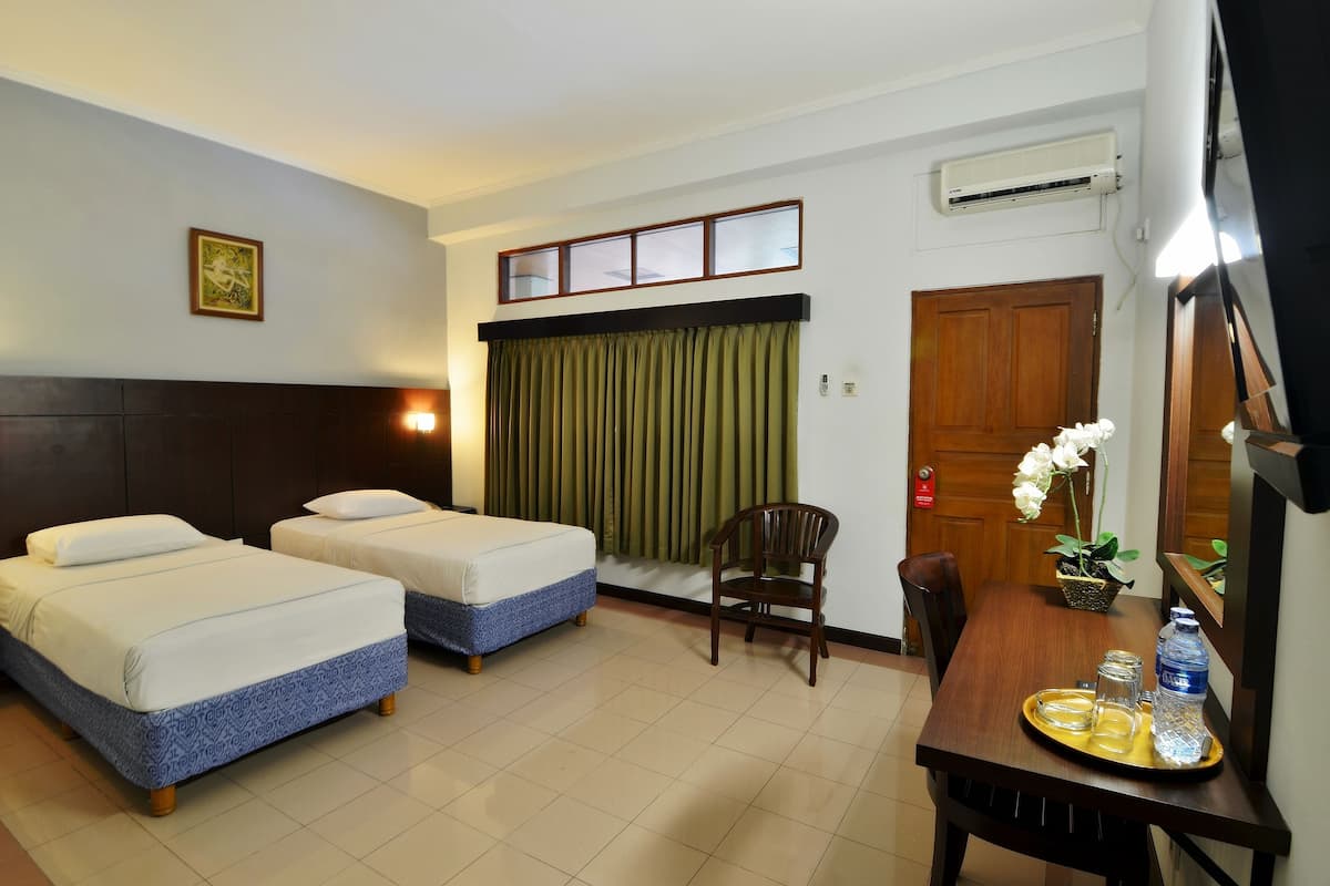 A hotel room with two beds, a dining table with a tray of glasses and a bottle, a chair, a potted plant, and an air conditioning unit.