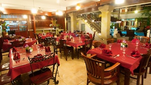 A restaurant with multiple tables set for dining, featuring red tablecloths and wooden chairs. - Rosani Hotel (Legian)