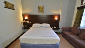 A hotel room with a large bed, a brown sofa, a nightstand with a phone, and a wall-mounted light fixture. - Rosani Hotel (Legian)