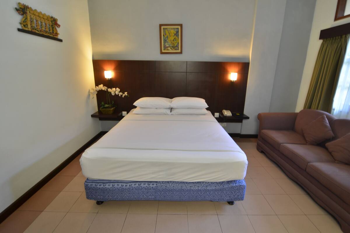 A hotel room with a large bed, a brown sofa, a nightstand with a phone, and a wall-mounted light fixture.