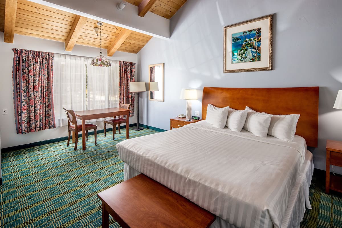 A hotel room with a large bed, wooden headboard, a dining table with chairs, a bench, and a large window with curtains.