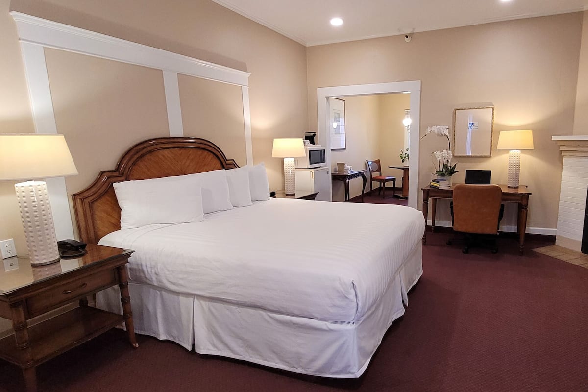 A hotel room with a large bed, bedside lamps, a desk with a chair, a fireplace, and a mirror on the wall.