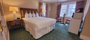 A hotel room with a large bed, a desk with a lamp, a chair, a small table with a plant, and a refrigerator.