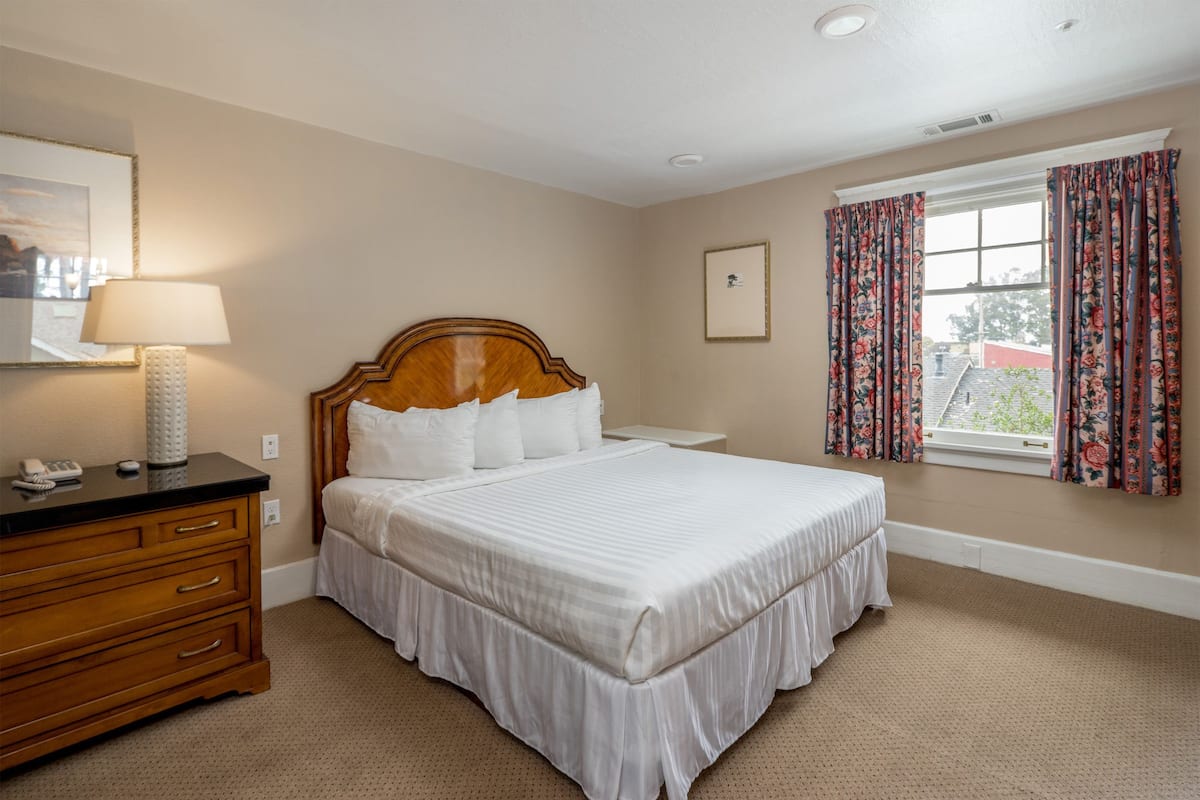 A neatly arranged bedroom with a large bed, a wooden dresser, a bedside lamp, and a window with floral curtains.