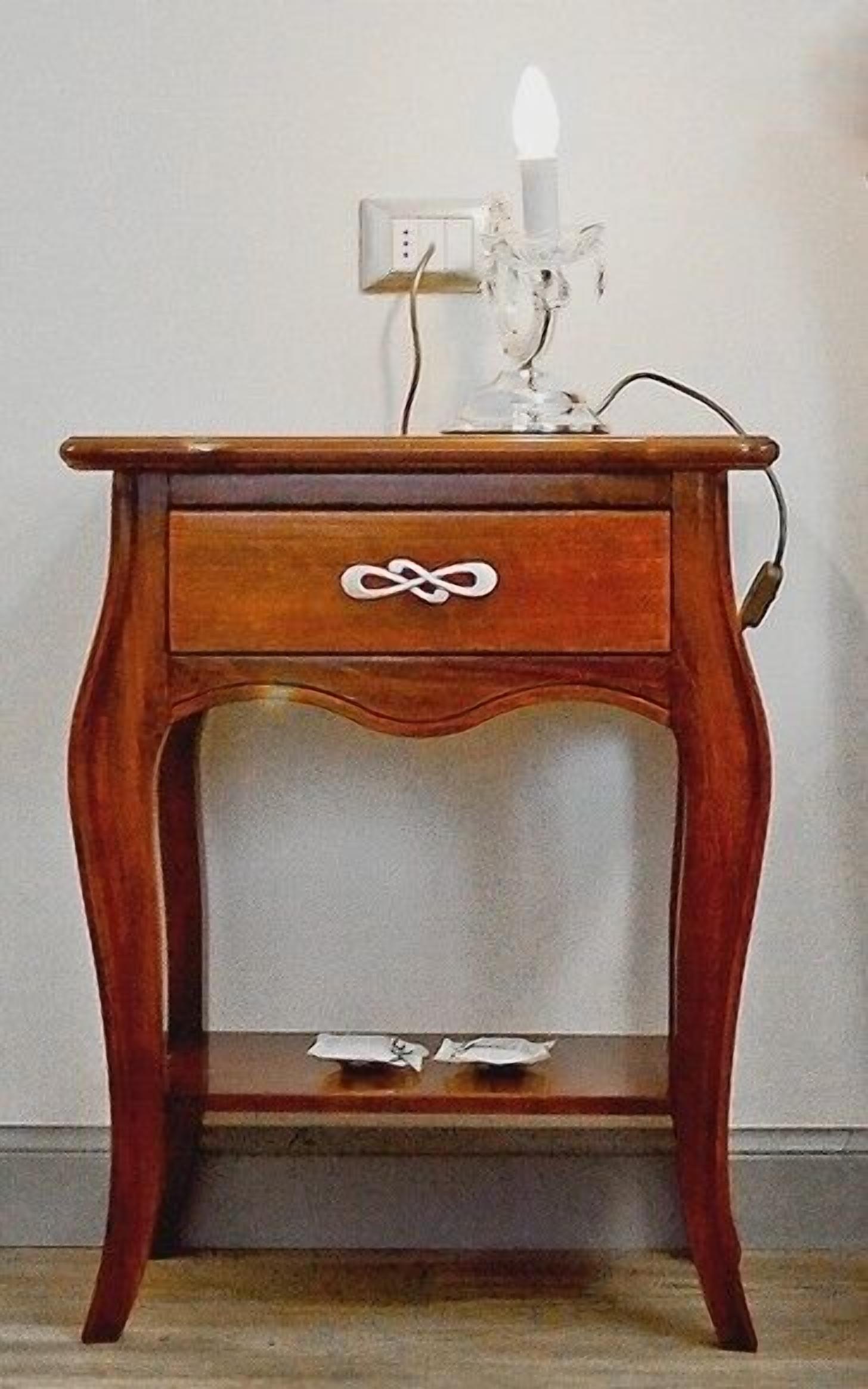 Una mesita de noche de madera con un cajón y un estante, un enchufe de pared y un candelabro.
