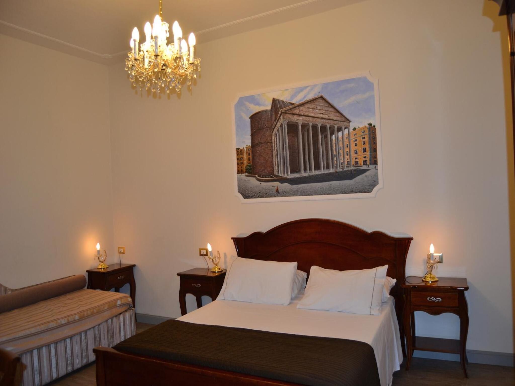 Quarto de hotel com uma cama grande, duas mesas de cabeceira com luminárias, um banco e um candelabro.