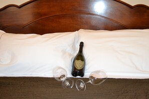 Un lit avec une tête de lit en bois, une bouteille de vin et deux verres à vin vides posés sur un drap blanc.