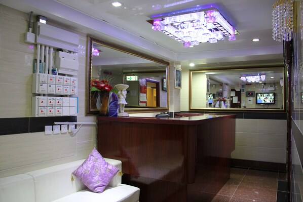 A hotel reception area with a wooden reception desk, a large mirror, and a white sofa with a purple pillow.