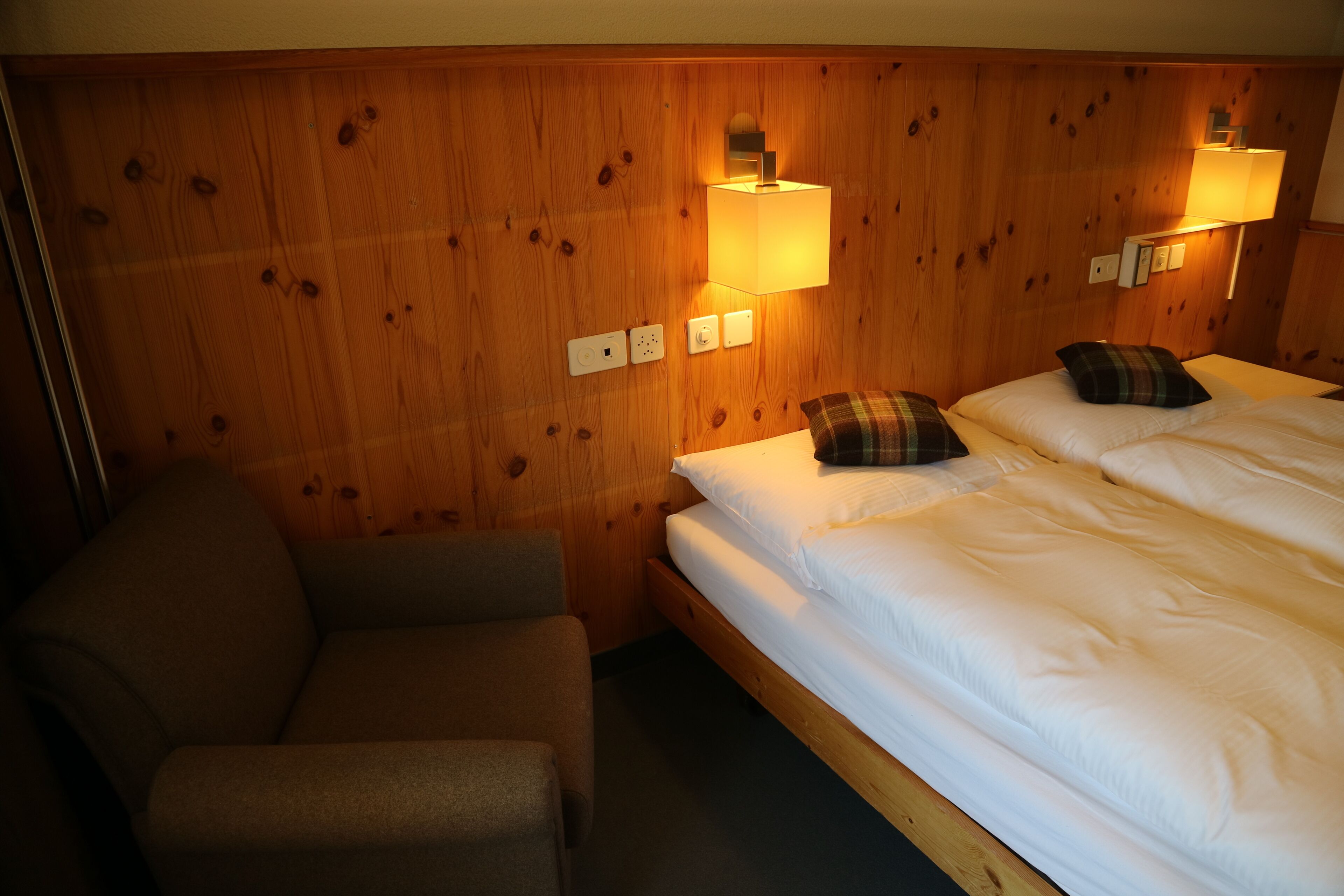 Une chambre d’hôtel avec une tête de lit en bois, deux lits avec du linge de lit blanc, un fauteuil marron et deux lampes de chevet.