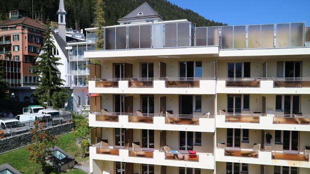 Um hotel com vários andares, varandas, uma igreja e montanhas ao fundo.