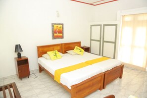 A neatly made bed with a wooden headboard, a bedside table with a lamp, a framed picture on the wall, and a window with curtains. - Ranveli Beach Resort (Mount Lavinia)