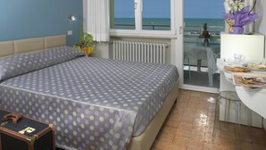 Hotelkamer met een bed, een klein tafeltje met stoel, een raam met uitzicht op zee en een koffer op de vloer.