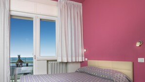 Een slaapkamer met een bed, nachtkastje, raam met gordijnen en uitzicht op zee.