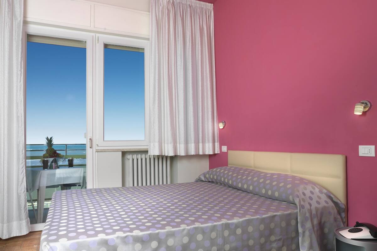 A bedroom with a bed, a nightstand, a window with curtains, and a view of the sea.
