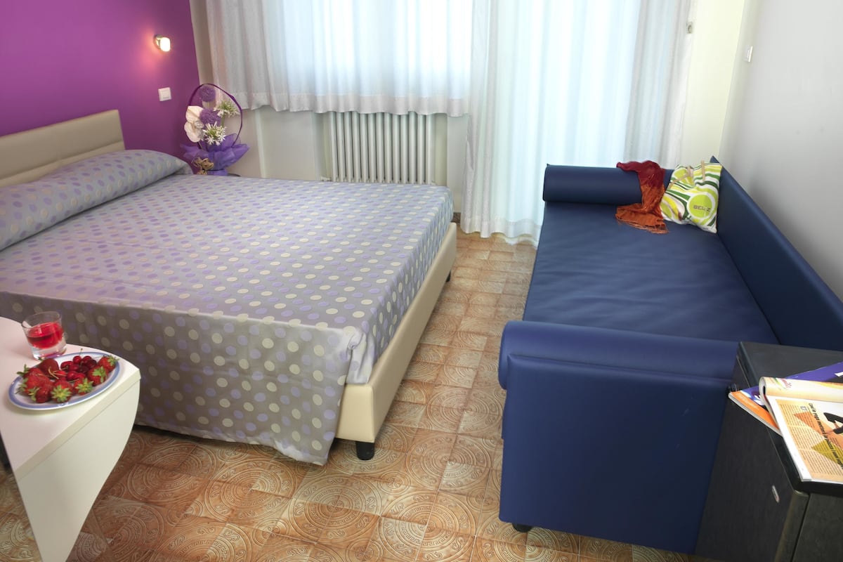 A room with a bed, a sofa, a small table with a plate of strawberries, and a window with curtains.