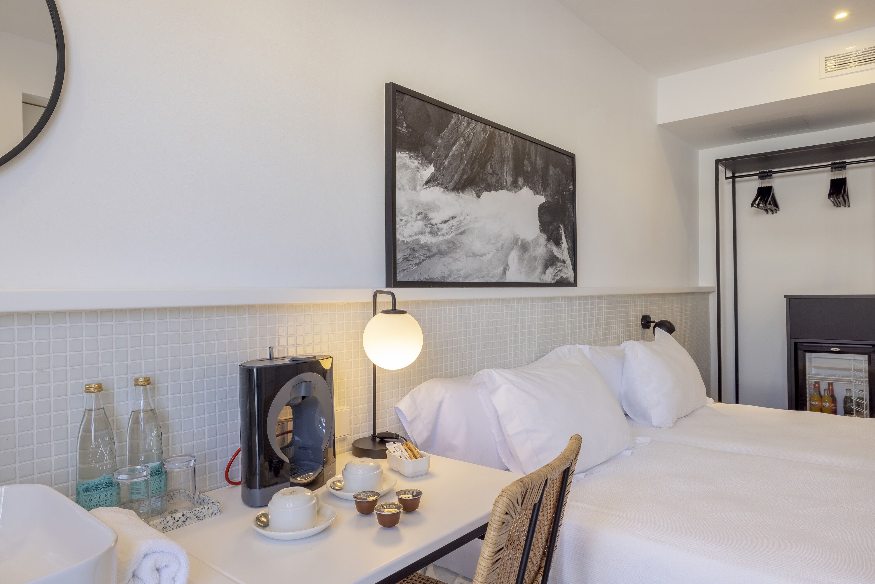 A hotel room with a bed, a desk with a coffee machine, a lamp, and a framed picture on the wall.