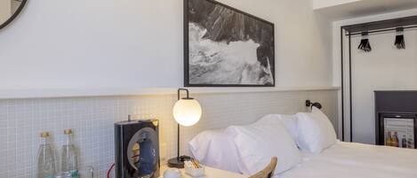 A hotel room with a bed, a desk with a coffee machine, a lamp, and a framed picture on the wall.