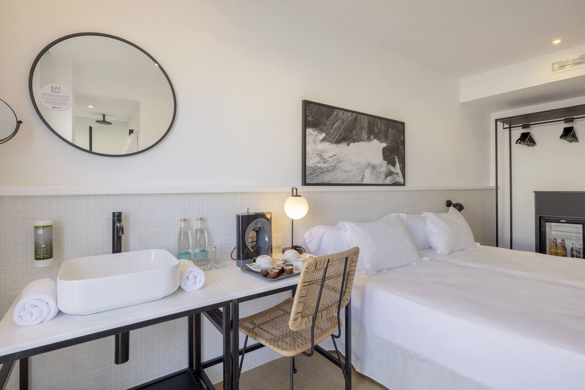 A hotel room with a bed, a desk with a sink, a chair, a round mirror, a wall-mounted picture, and a wardrobe with clothes.
