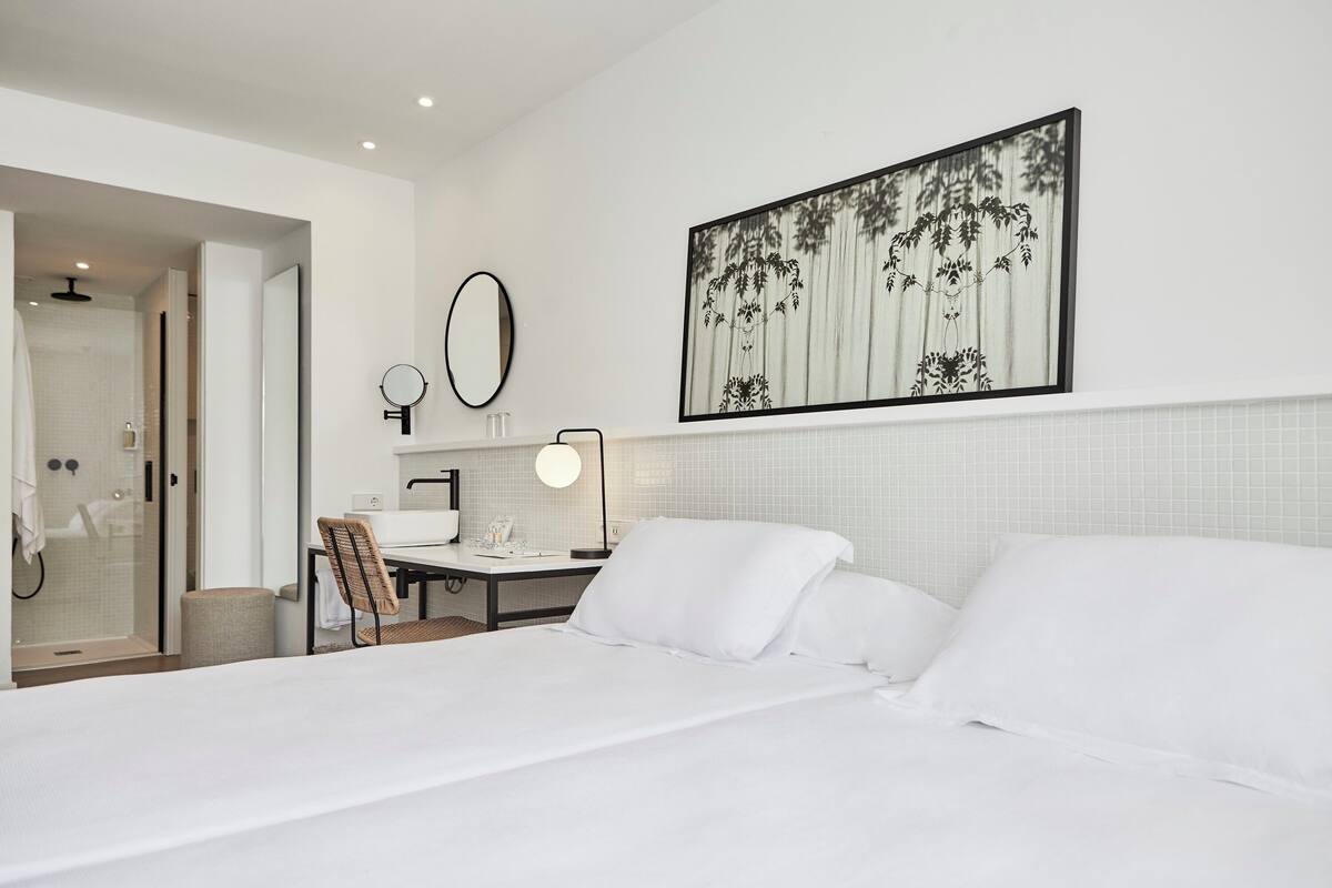 A modern hotel room with a large bed, a desk with a chair, a mirror, and a wall-mounted picture.