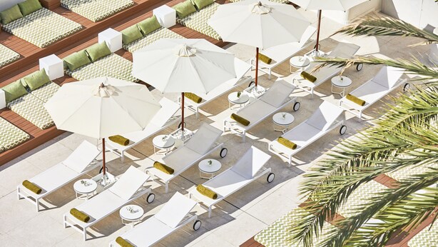 An outdoor seating area with white lounge chairs and tables, arranged in rows with umbrellas providing shade.