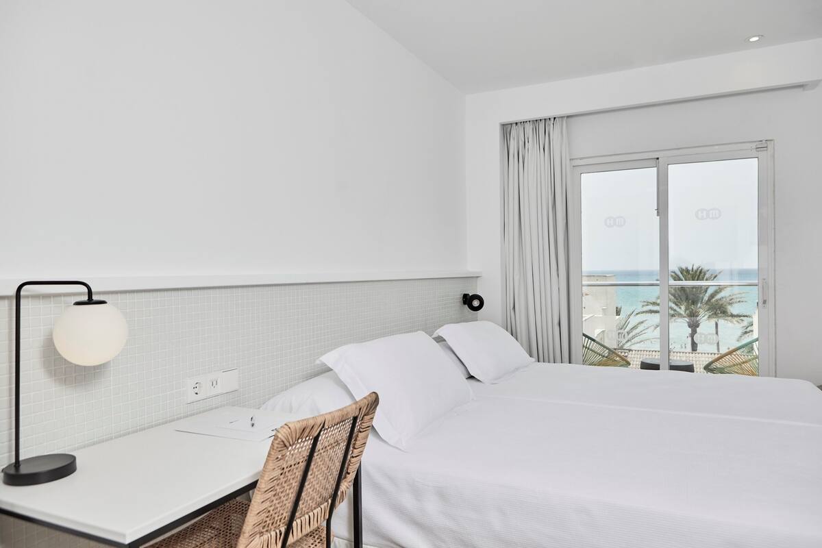 A hotel room with a bed, a desk with a lamp, a chair, and a view of the ocean through a large window.