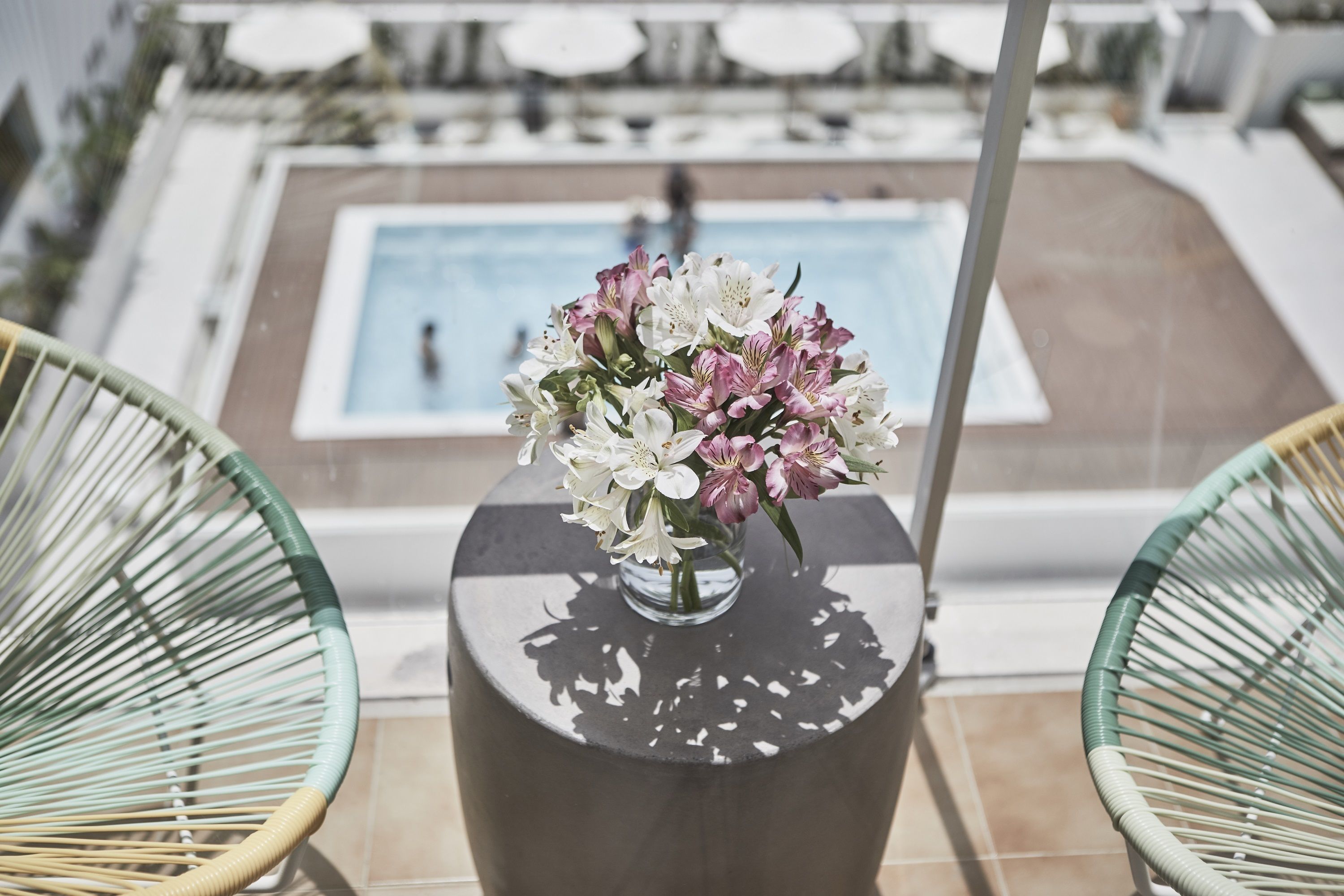 Zona de piscina con una mesa redonda que sostiene un jarrón de flores, dos sillas de mimbre y una piscina al fondo.