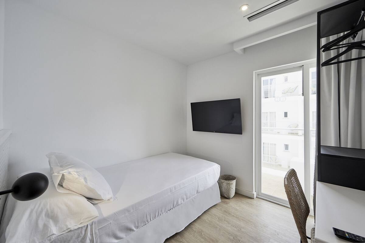 A minimalist bedroom with a bed, a TV mounted on the wall, and a view of the outside through a sliding door.