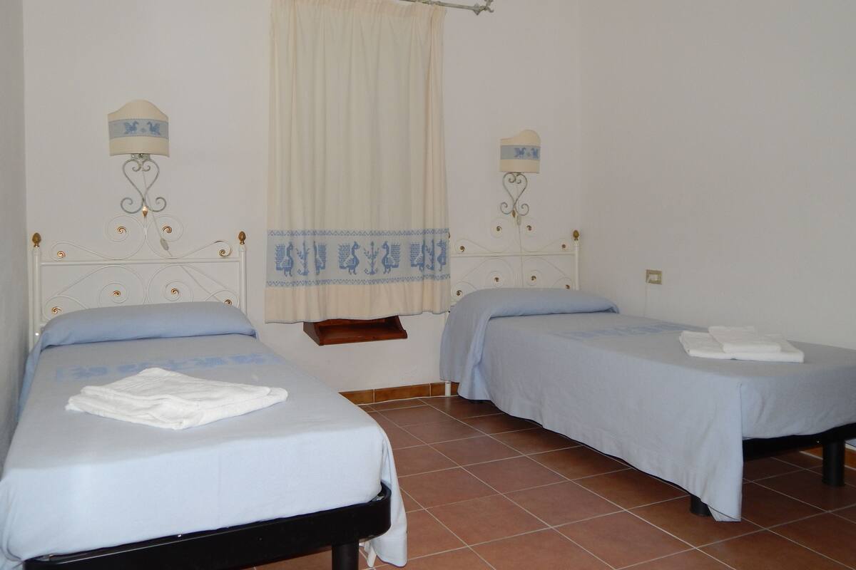 A room with two single beds, each with a white mattress and a blue blanket. There is a window with a curtain, and wall-mounted lamps on either side of the window.