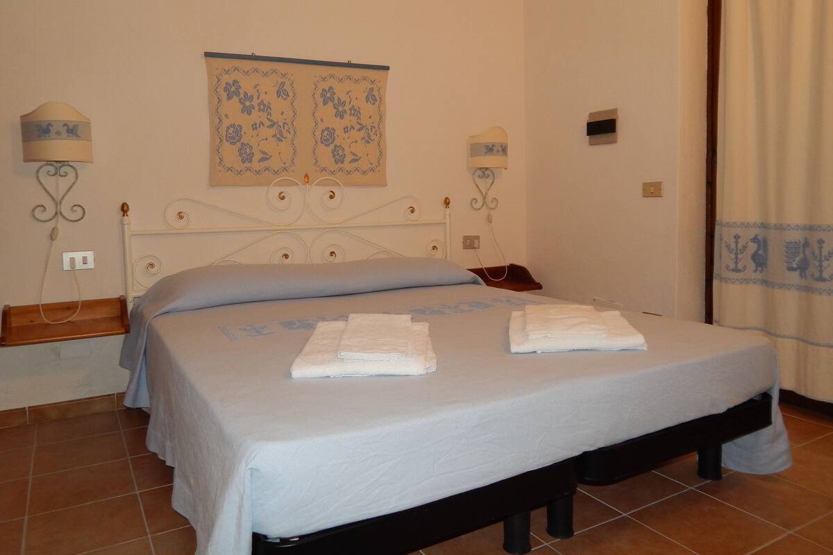 A neatly made bed with two folded towels, a wall-mounted lamp, and a decorative tapestry.