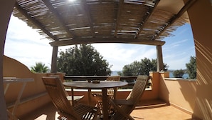Une terrasse extérieure couverte, avec une table ronde et quatre chaises en bois, offrant une vue panoramique sur un paysage arboré et un plan d’eau.