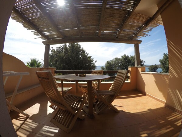 A covered outdoor patio with a round table and four wooden chairs, overlooking a scenic view of trees and a body of water.