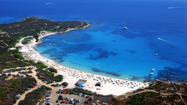 Vue aérienne d’une zone côtière avec une plage, des bateaux et des voitures garées.