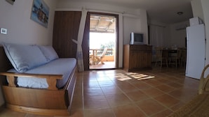 Une chambre avec un lit en bois, un téléviseur posé sur un meuble en bois, un coin repas avec des chaises et une table, et une vue sur l’extérieur.