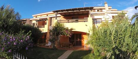 Un edificio de dos plantas con techo de tejas, fachada de piedra y un balcón. Un camino conduce a la entrada, rodeado de follaje y una valla pequeña.