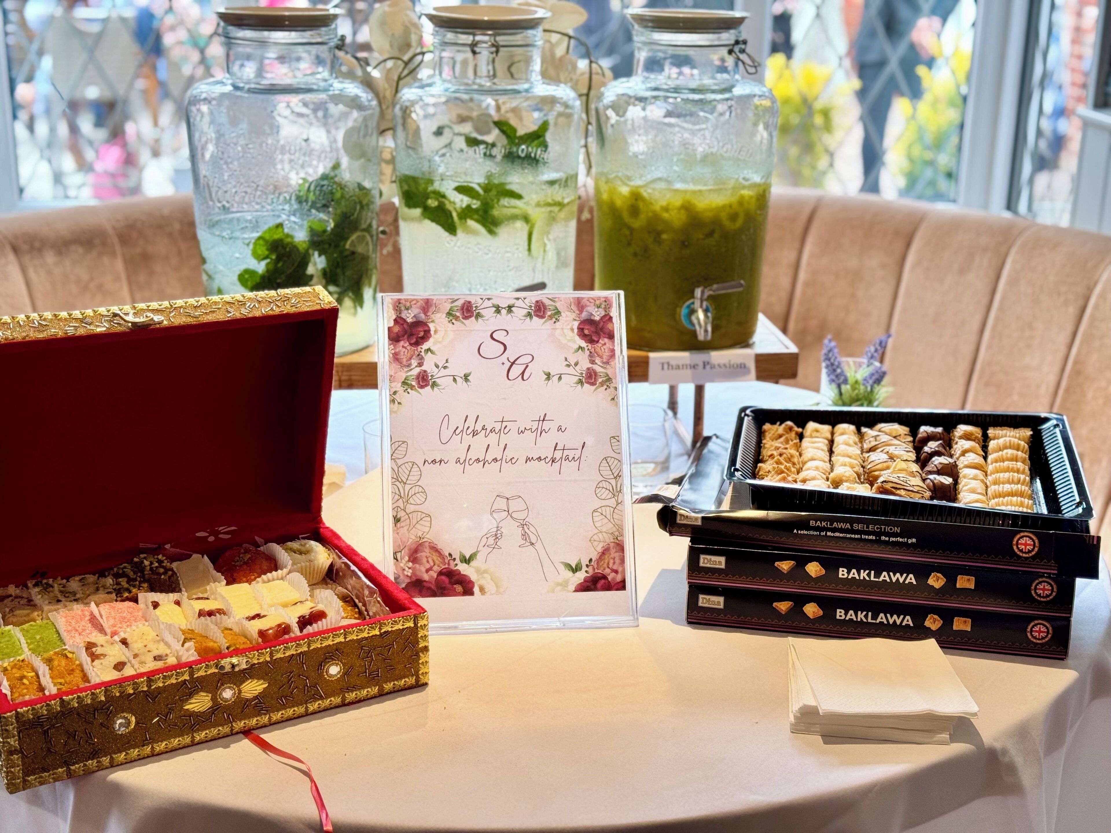 Een tafel met een keuze aan desserts, waaronder een rode velvet doos, een bord met een boodschap over een alcoholvrije cocktail, en potten vol drank.