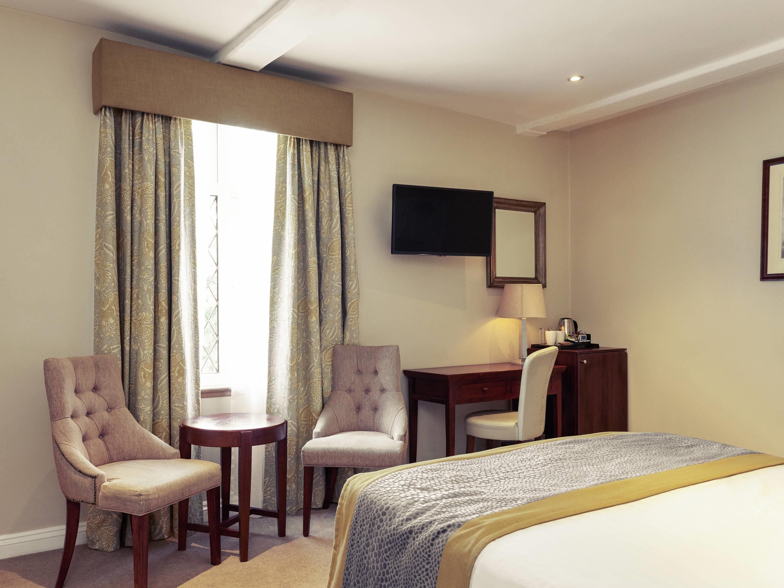 A hotel room with a bed, two armchairs, a desk, a lamp, and a television.