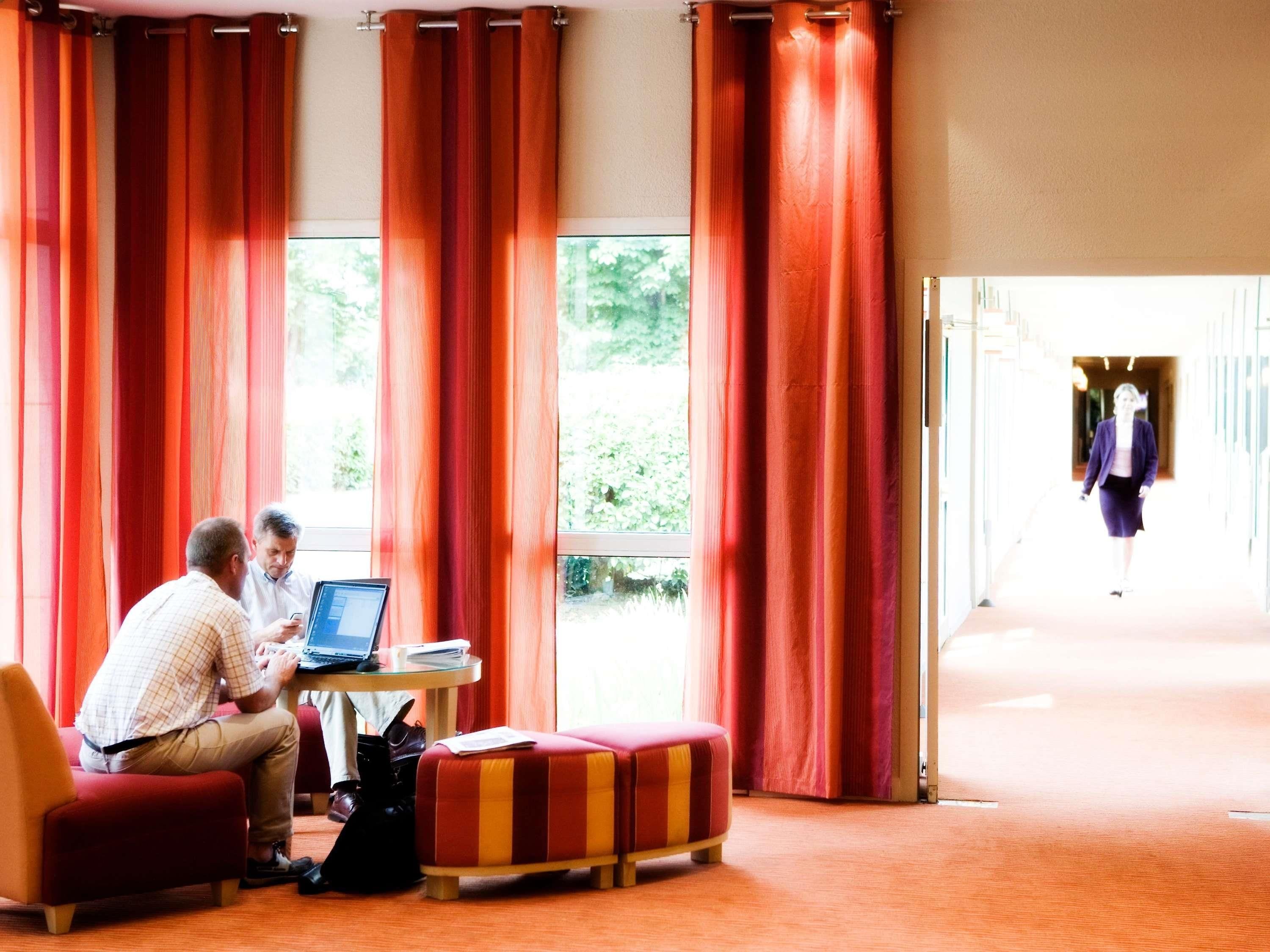 Une chambre d’hôtel avec de grandes fenêtres, des rideaux rouges, une petite table ronde avec un ordinateur portable, et un pouf rayé de rouge et de jaune.