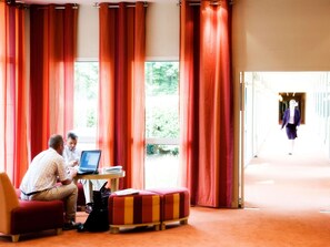 A hotel room with large windows, red curtains, a small round table with a laptop, and a red and yellow striped ottoman. - Mercure Thame Lambert Hotel (Watlington)