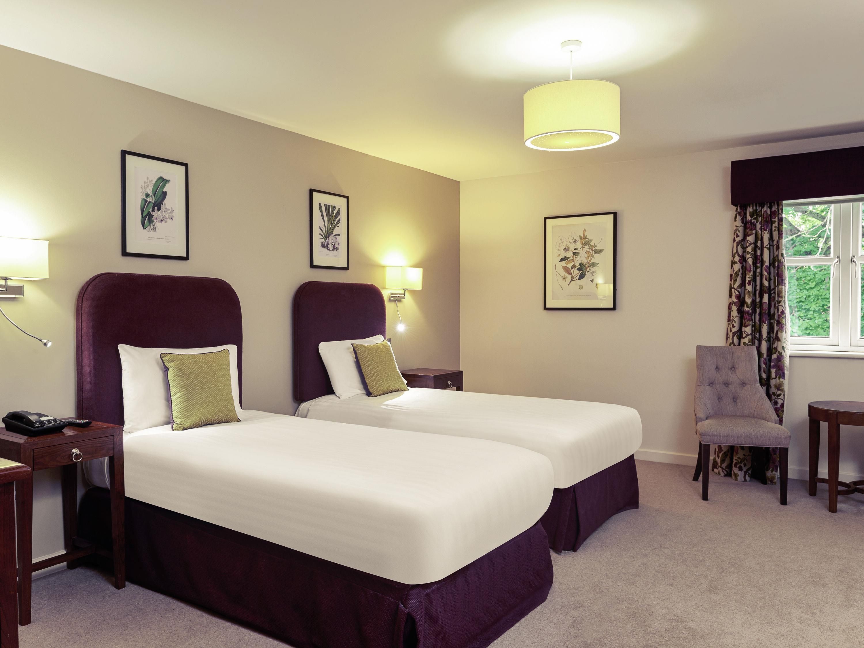 A hotel room with two beds, a chair, a desk, and framed botanical prints on the wall.