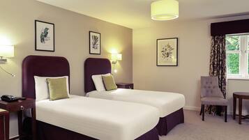 Ein Hotelzimmer mit zwei Betten, einem Sessel, einem Schreibtisch und gerahmten Pflanzenbildern an der Wand.