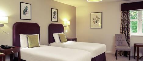 A hotel room with two beds, a chair, a desk, and framed botanical prints on the wall.