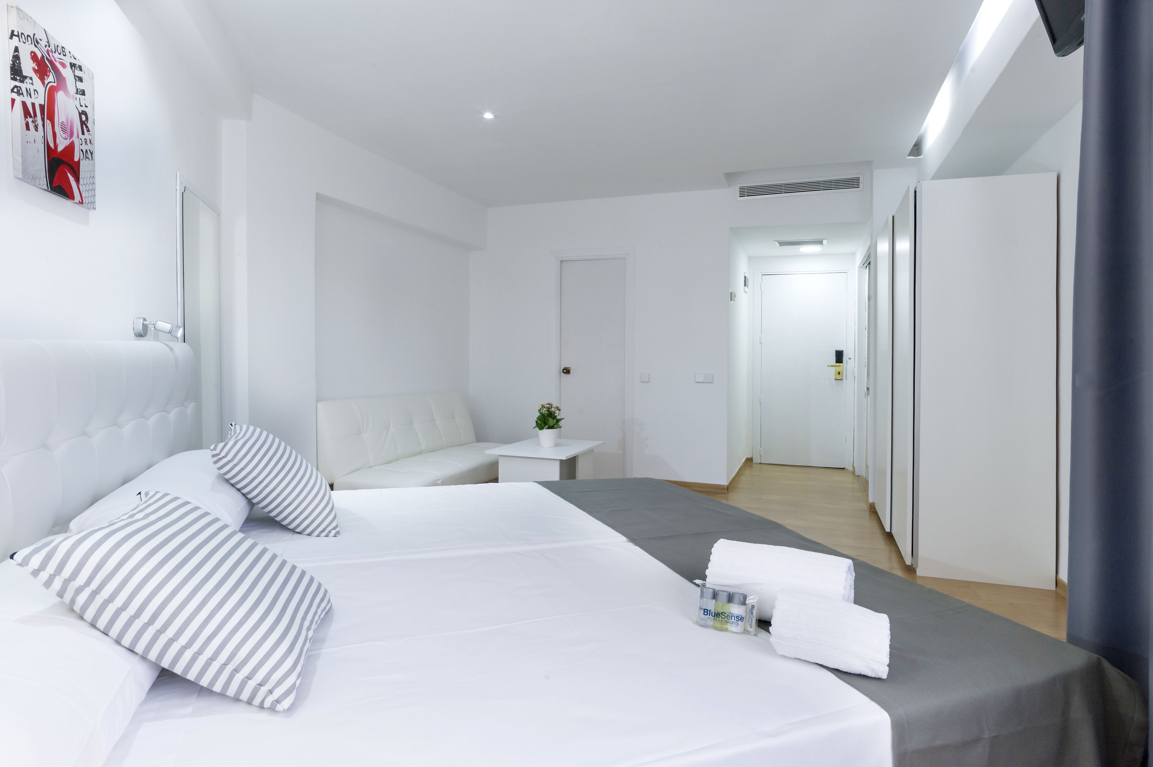 A hotel room with a bed, a small table with a plant, and a hallway leading to a door.