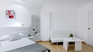 Ein modernes, minimalistisches Schlafzimmer mit einem großen Bett, einem Nachttisch mit Pflanze, einem weißen Sofa und einem kleinen Tisch.