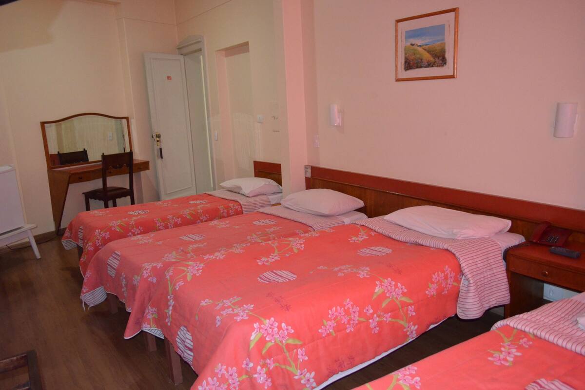Une chambre d’hôtel avec deux lits, une chaise en bois, un bureau, un miroir, une armoire et un tableau au mur.