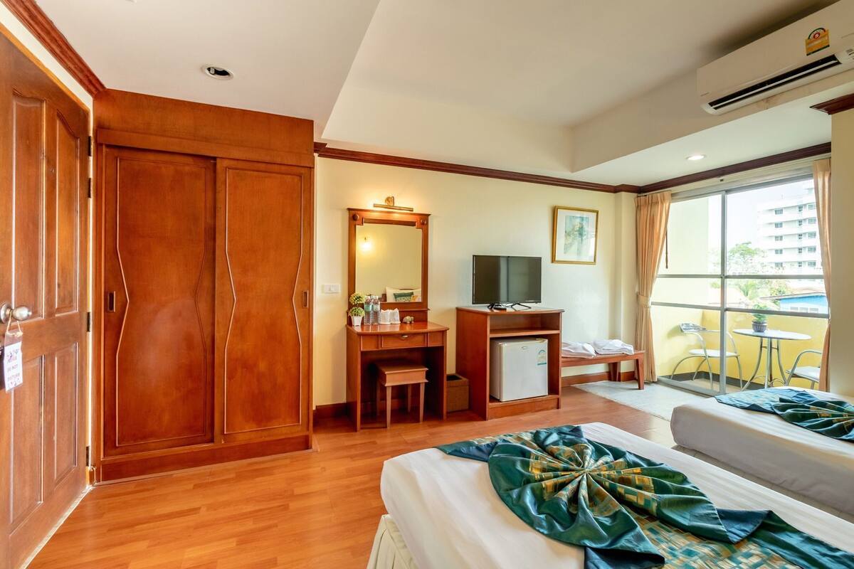 A hotel room with a wooden wardrobe, a desk with a mirror, a television, a bed with a green patterned bedspread, and a balcony with chairs and a table.