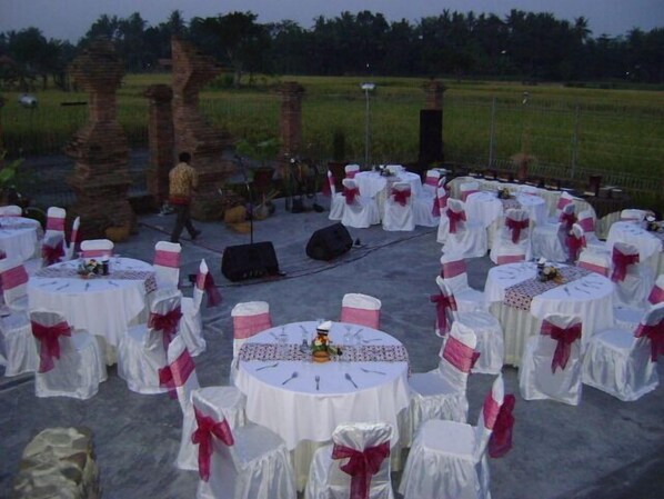 An outdoor event setup with round tables covered in white tablecloths and chairs with pink sashes, arranged in a semi-circle. - Tembi Rumah Budaya (Sewon)