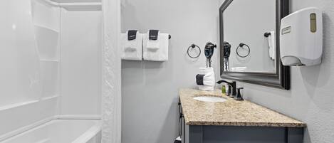 A modern bathroom with a bathtub, a sink with a granite countertop, a mirror, and a hand dryer.