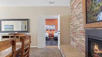 A room with a fireplace, a dining area with wooden furniture, and a view into a bedroom with a bed and a nightstand.