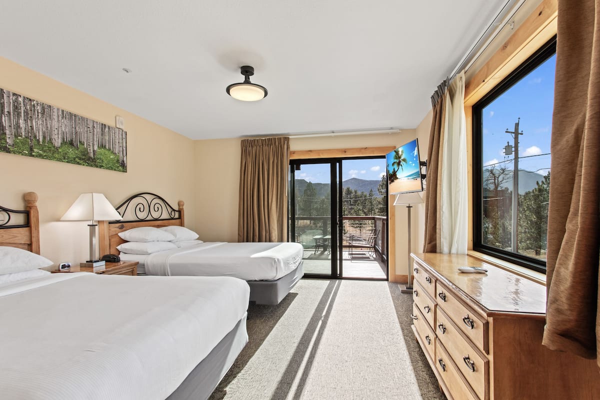 Habitación de hotel con dos camas, un aparador, un televisor y un balcón con vista a las montañas.